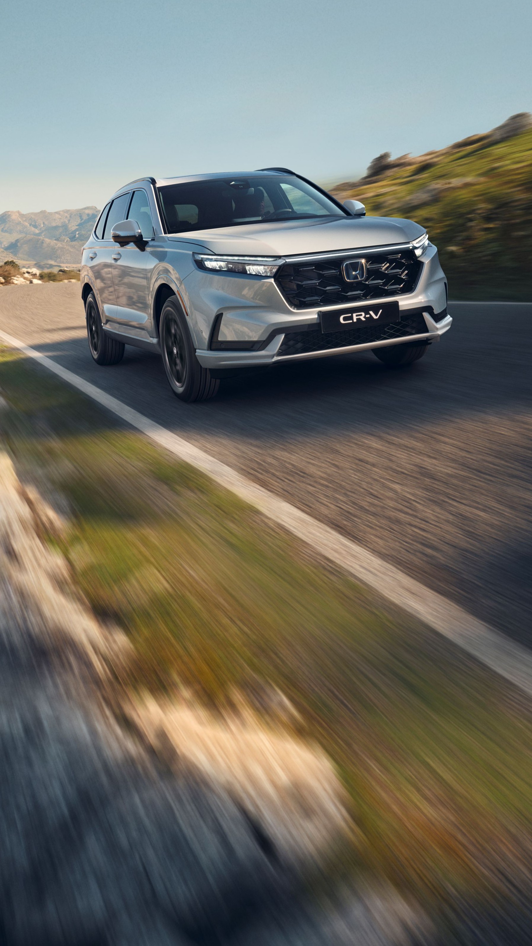 Un SUV Honda CR-V argento in marcia su una strada di montagna tortuosa con terreno roccioso e montagne sullo sfondo. Il veicolo è in movimento, creando una sensazione di velocità e avventura nel paesaggio impervio.