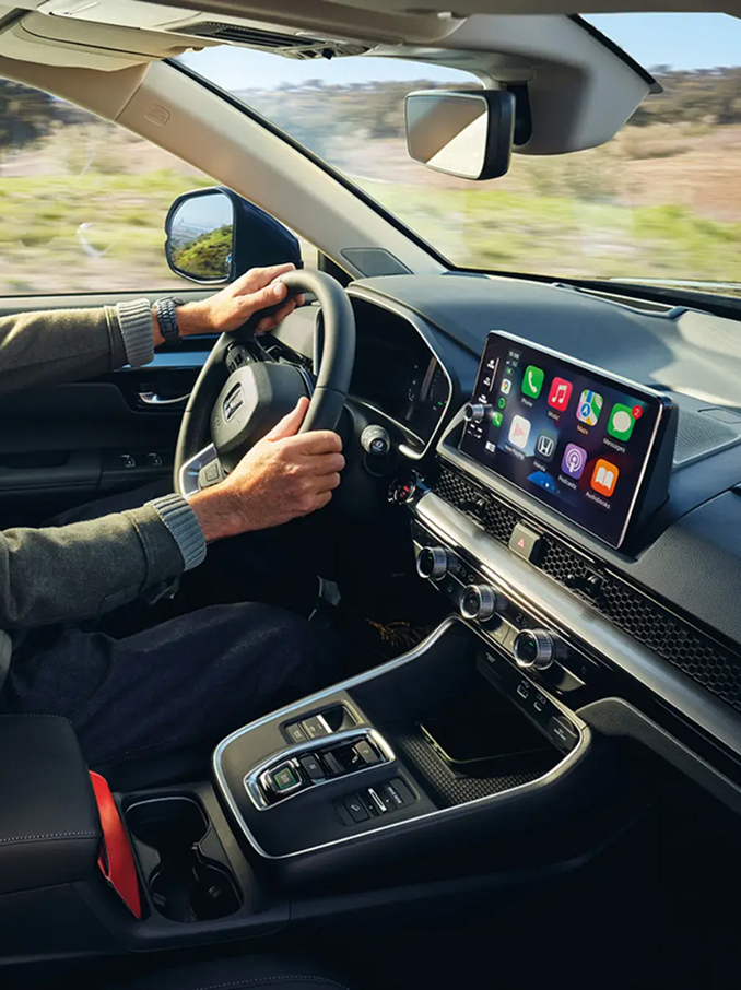 Vista interna di un'auto moderna con il conducente seduto al volante, che mostra la plancia, il volante, la console centrale e un ampio display touchscreen con icone di app colorate, mentre un paesaggio rurale scorre fuori dai finestrini.