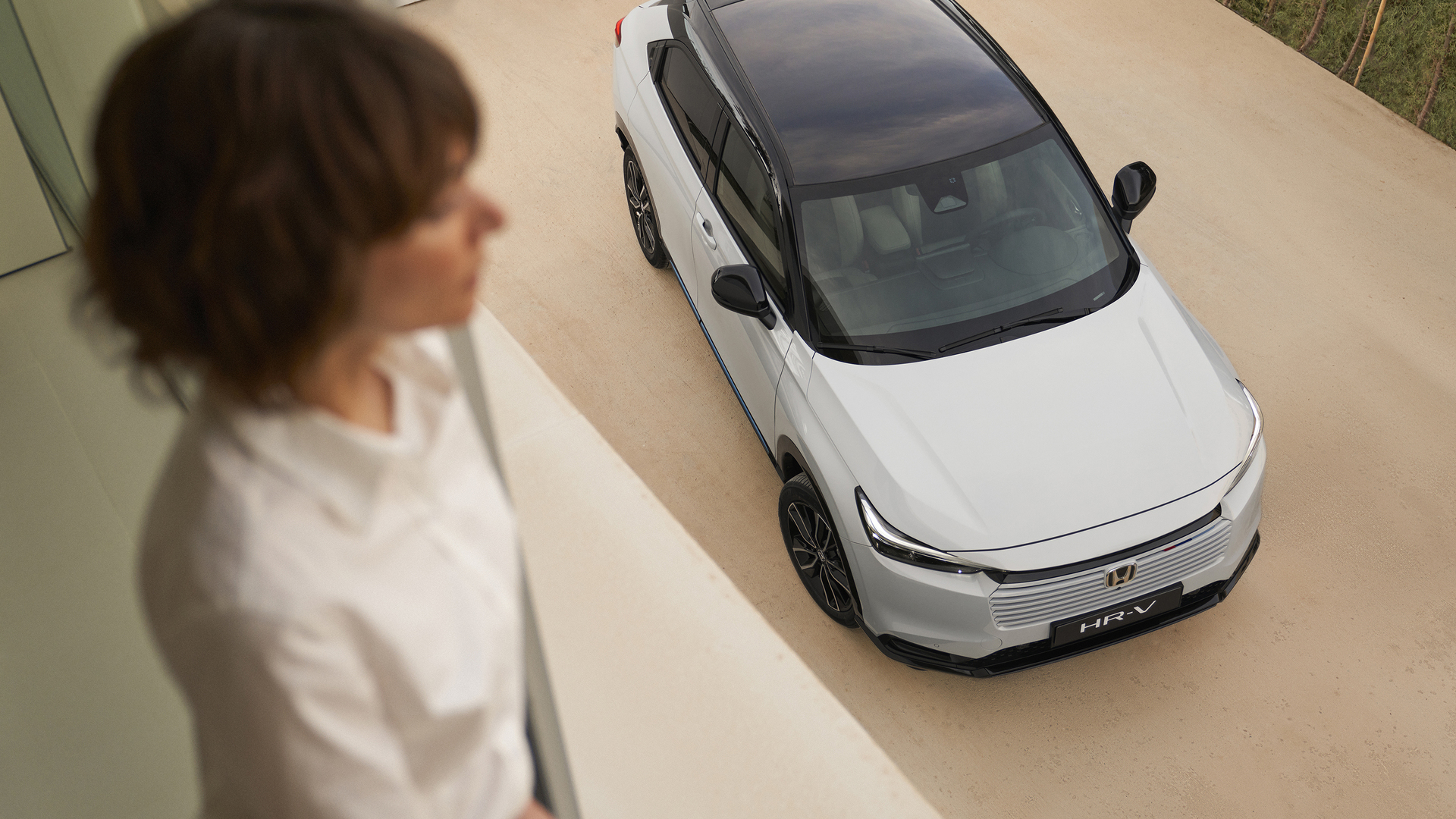 Vista dall'alto di una Honda HR-V bianca con tetto nero parcheggiata su un vialetto chiaro, mentre una persona sfocata si trova all'interno in primo piano e guarda verso l'auto.