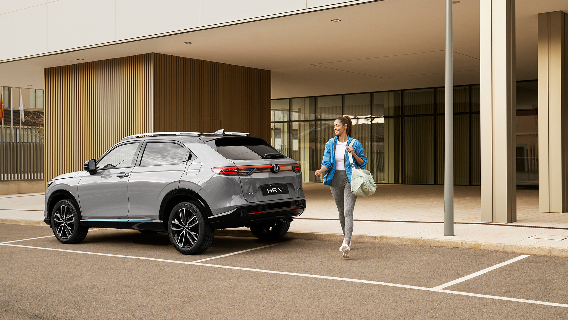 Una Honda HR-V color argento è parcheggiata davanti a un edificio moderno mentre una donna passa accanto portando una borsa, suggerendo un utilizzo urbano quotidiano e praticità.