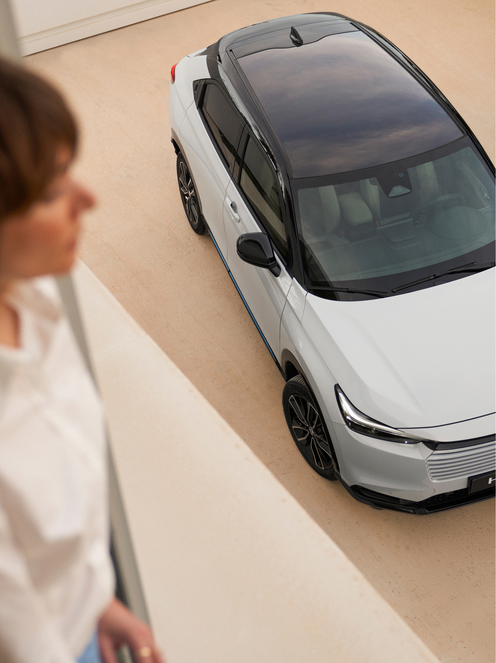 Vista dall'alto di una Honda HR-V bianca con tetto nero parcheggiata su un vialetto chiaro, mentre una persona sfocata si trova all'interno in primo piano e guarda verso l'auto.