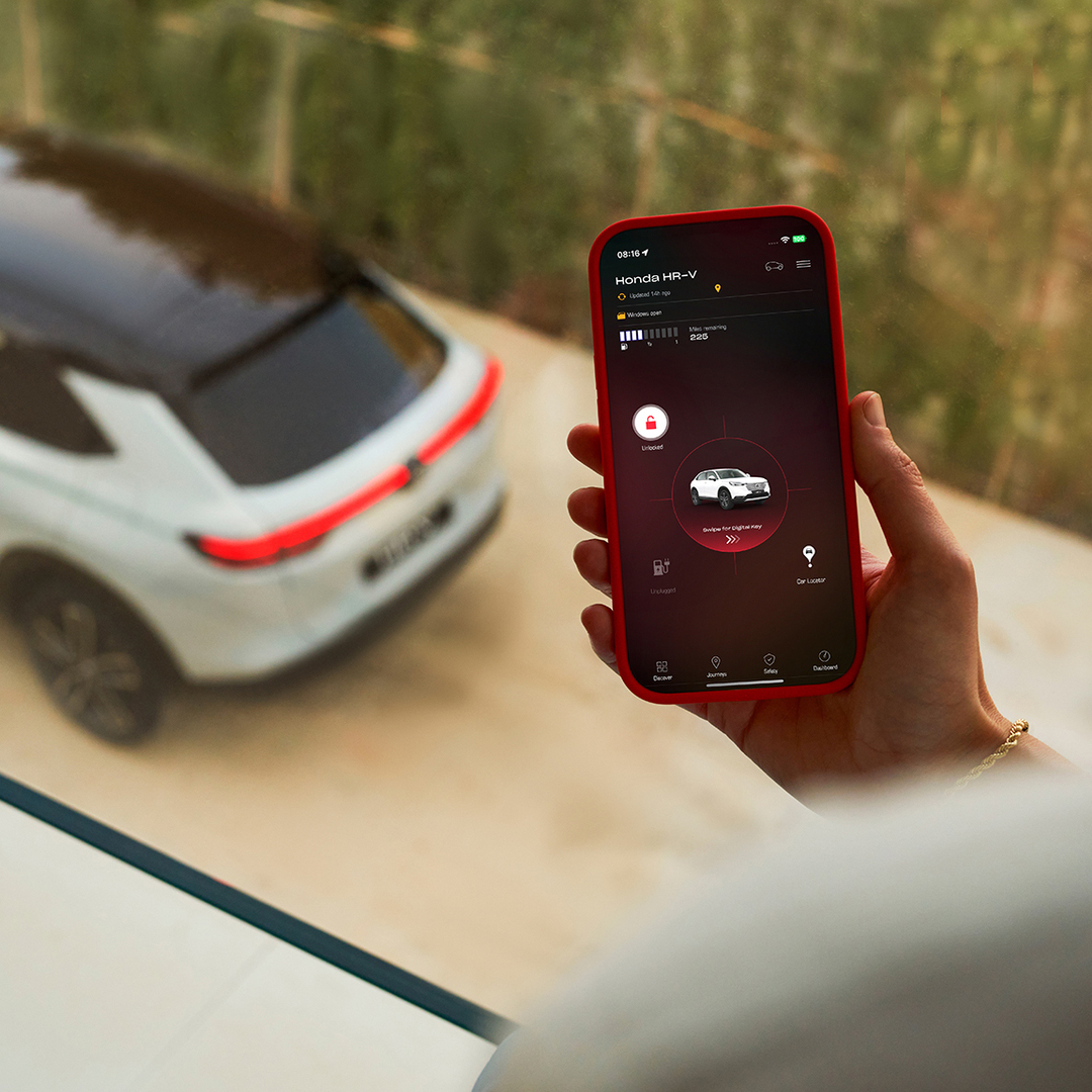Una persona tiene in mano uno smartphone che mostra un'app per le impostazioni di ricarica del veicolo, mentre sullo sfondo è parcheggiato un SUV bianco.
