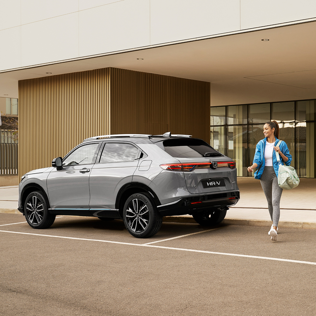 Una Honda HR-V color argento è parcheggiata davanti a un edificio moderno mentre una donna passa accanto portando una borsa, suggerendo un utilizzo urbano quotidiano e praticità.