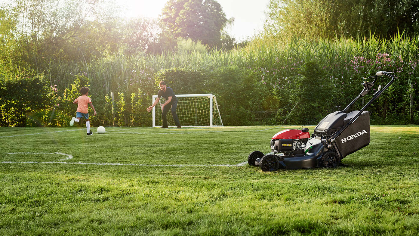 Uomo e bambino che giocano a calcio accanto a Honda HRN visto di fianco in giardino. 