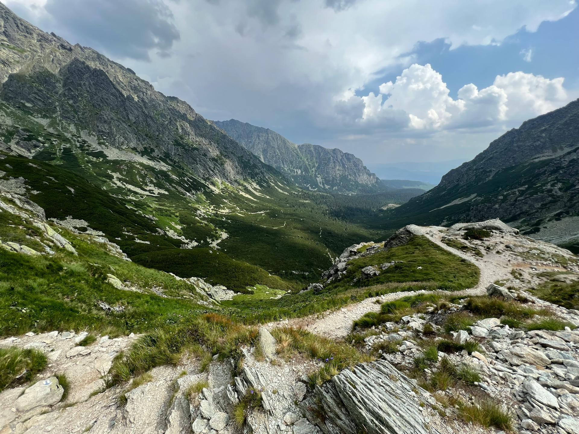 A scenic mountain landscape with a winding dirt path leading through a rocky valley. Steep, rugged peaks flank both sides of the green valley floor. In the distance, the valley opens up to a hazy horizon under a partly cloudy sky.