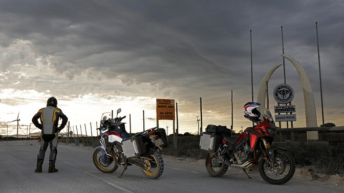 Guidatore che guarda l'orizzonte in piedi accanto a due Africa Twin parcheggiate sull'asfalto.