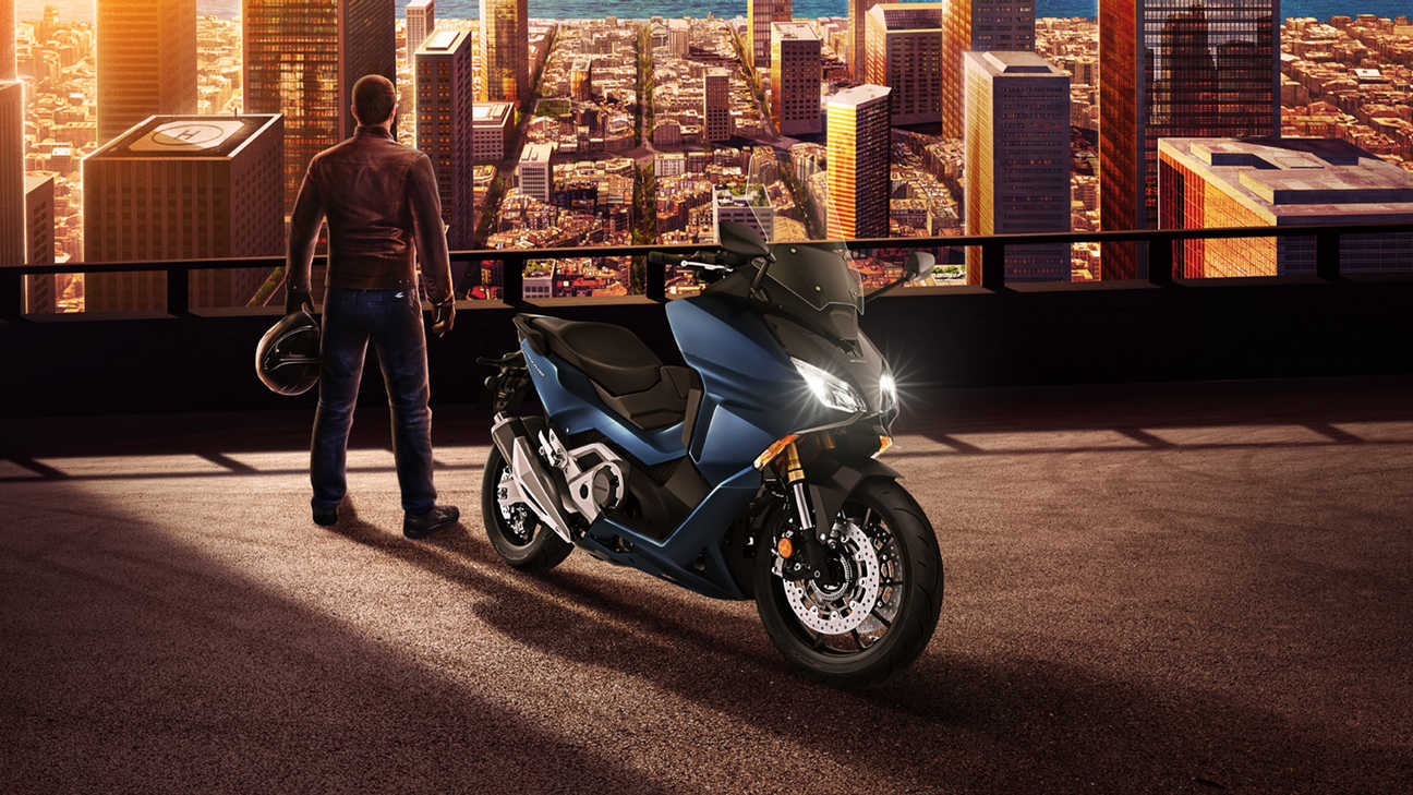 Honda Forza 750, vista anteriore di tre quarti lato destro, uomo in piedi accanto alla moto parcheggiata che sovrasta la città, moto blu