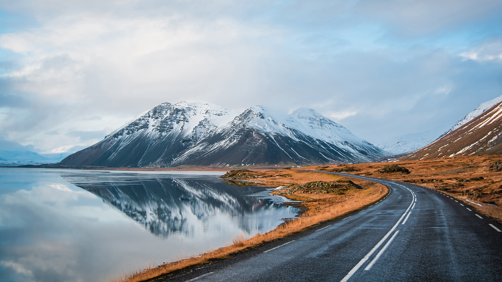 Foto panoramica invernale della strada che conduce lungo la costa del lago fino alle montagne vulcaniche. Alte vette rocciose ricoperte da uno strato di neve che si specchia sulla superficie dell'acqua. Punto di vista del conducente sulla strada circolare, Islanda.