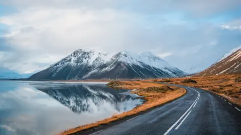 Foto panoramica invernale della strada che conduce lungo la costa del lago fino alle montagne vulcaniche. Alte vette rocciose ricoperte da uno strato di neve che si specchia sulla superficie dell'acqua. Punto di vista del conducente sulla strada circolare, Islanda.