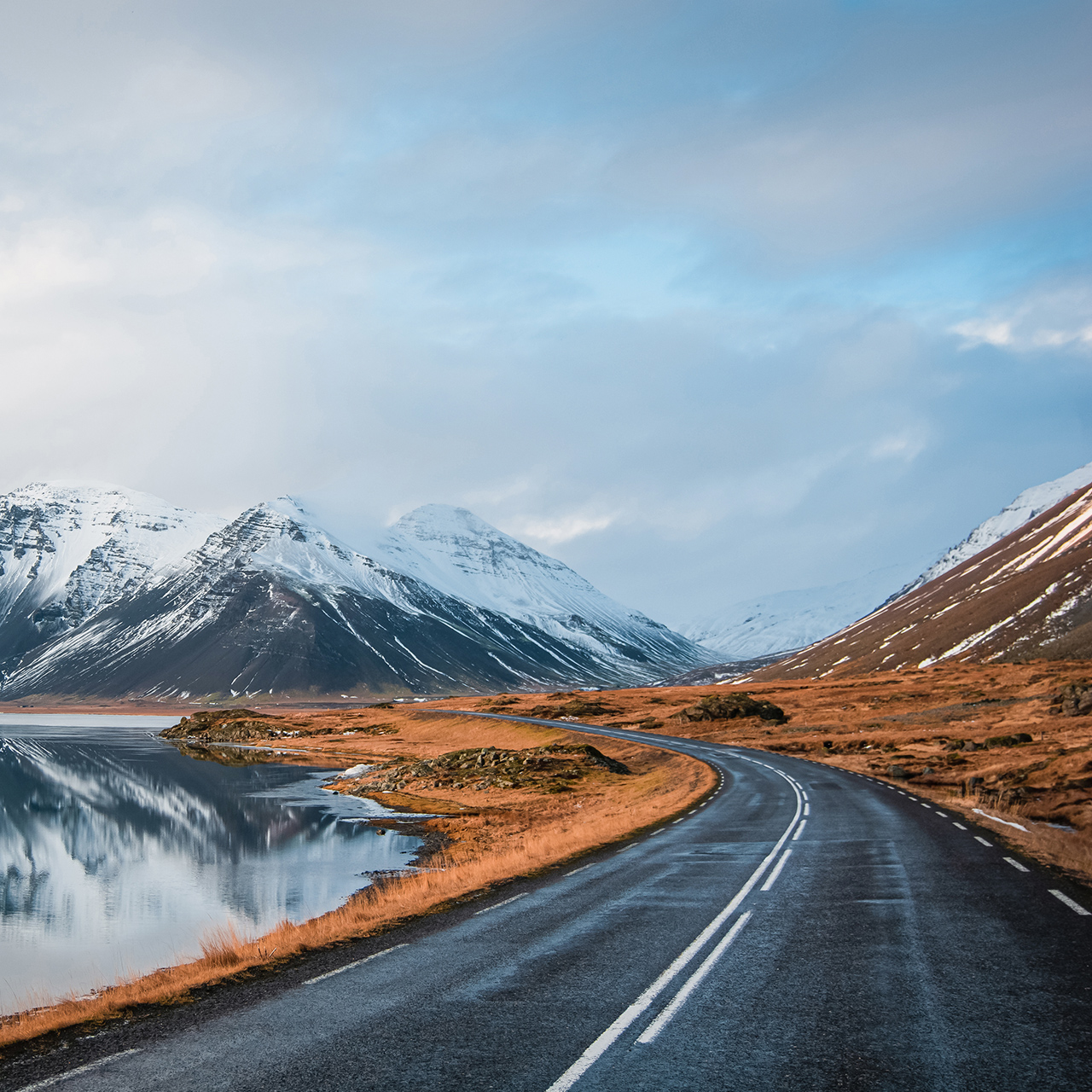 Foto panoramica invernale della strada che conduce lungo la costa del lago fino alle montagne vulcaniche. Alte vette rocciose ricoperte da uno strato di neve che si specchia sulla superficie dell'acqua. Punto di vista del conducente sulla strada circolare, Islanda.
