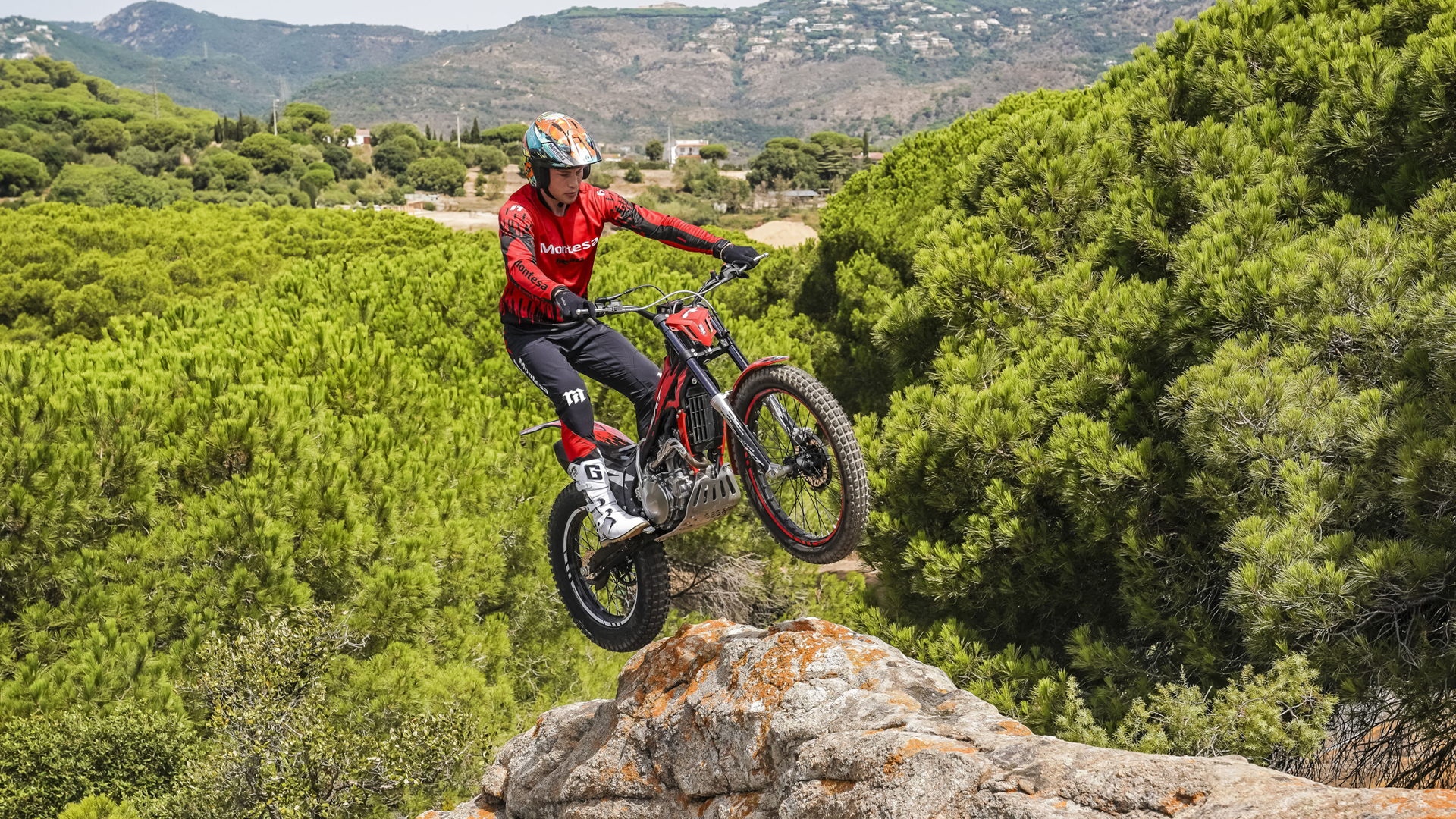 Un motociclista salta da una sporgenza rocciosa sopra una pineta panoramica con Montesa Cota 4RT 260R rossa.