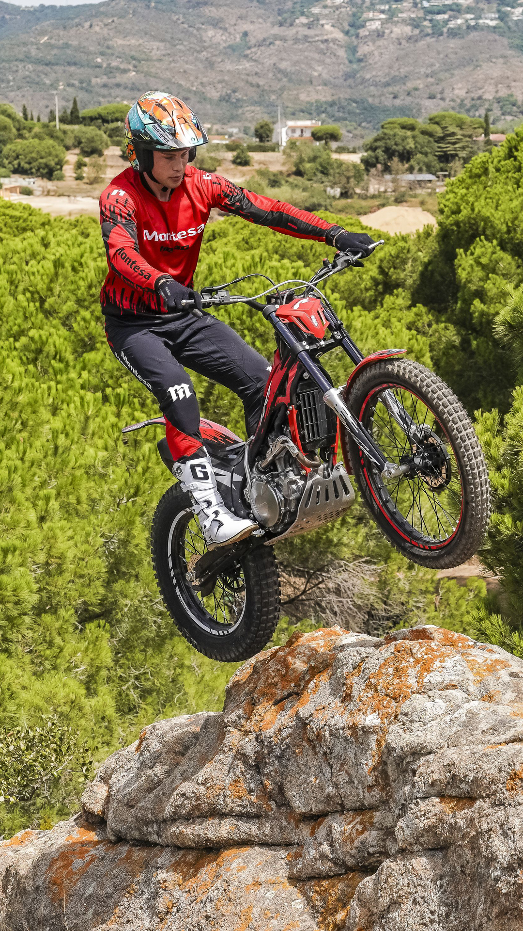 Un motociclista salta da una sporgenza rocciosa sopra una pineta panoramica con Montesa Cota 4RT 260R rossa.