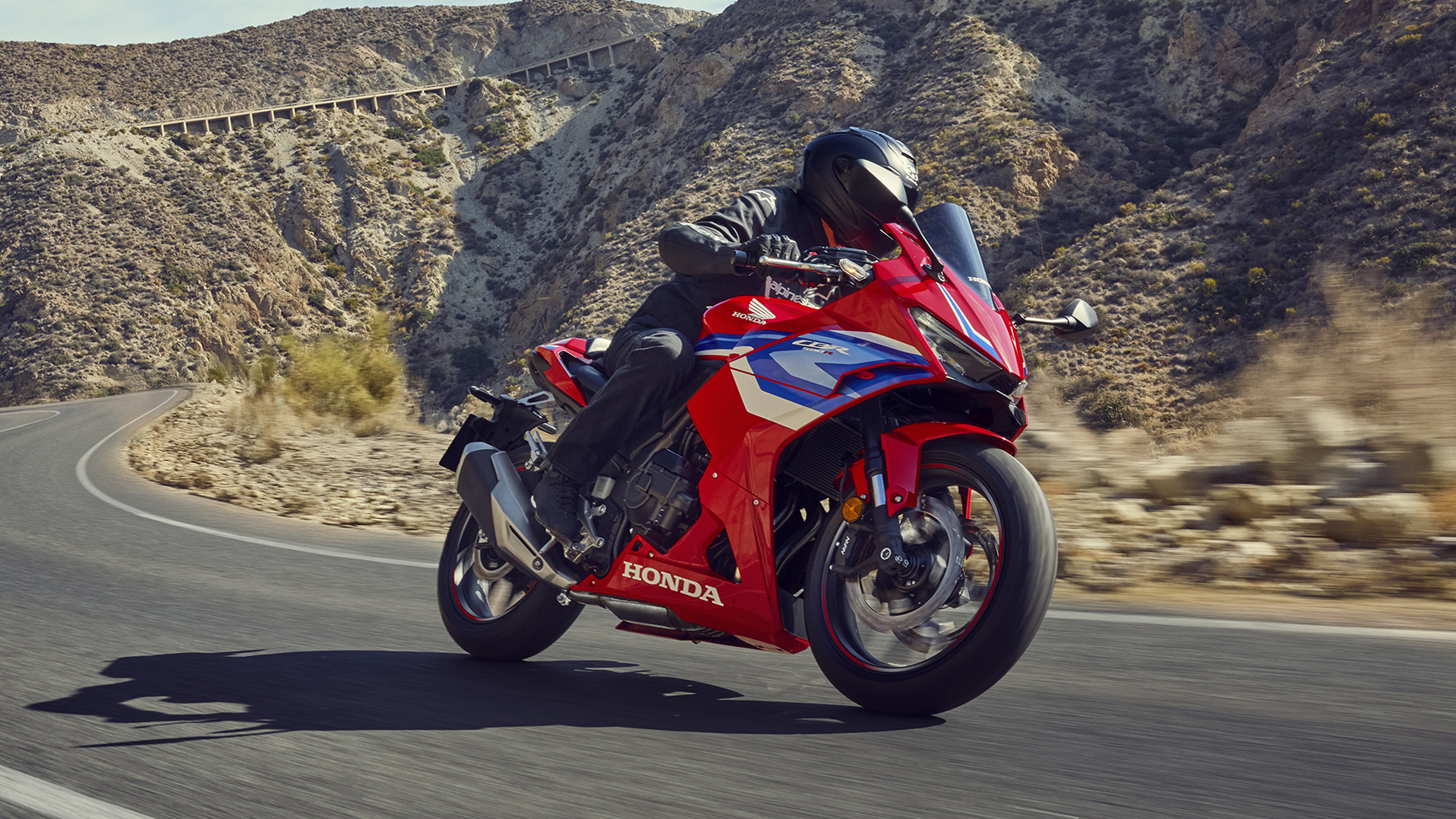 A red Honda CBR500R motorcycle rides on a winding mountain road. The sport bike is in motion, leaning into a curve, with rocky, arid hills and a distant bridge visible in the background.