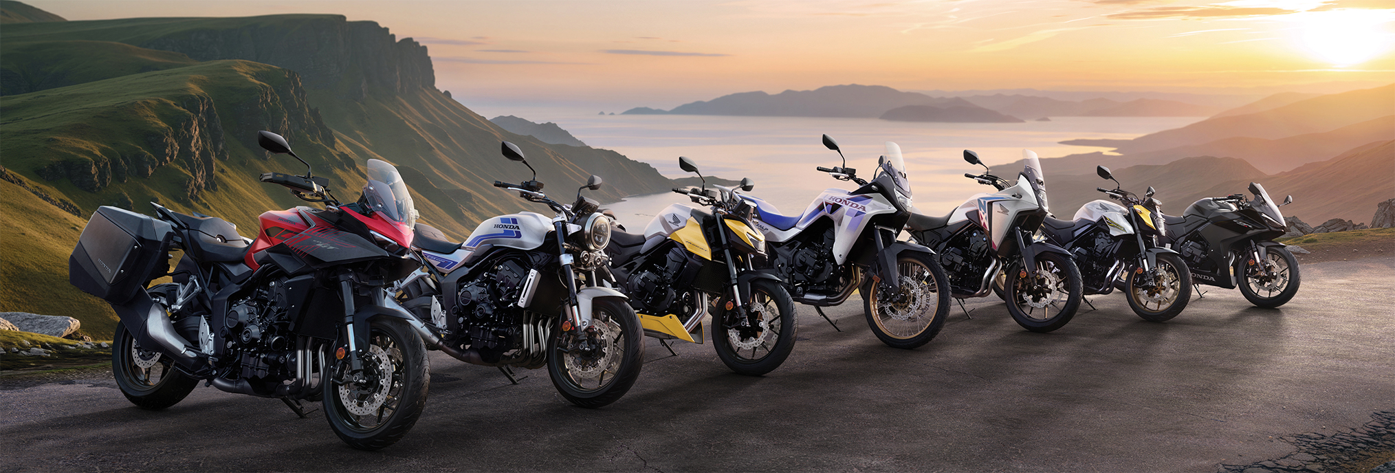 A lineup of seven Honda motorcycles parked on a scenic mountain road at sunset. The bikes range from sport to adventure models, showcasing the diverse Honda motorcycle lineup against a backdrop of rolling hills and a golden sky.